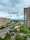 A cityscape featuring several high-rise apartment buildings on either side of a wide road. The area is lush with greenery, including trees lining the streets and a patch of cultivated land. There is notable construction work on one of the taller buildings. The sky is cloudy, suggesting an overcast day.