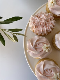 A few cupcakes with light pink frosting are arranged on a white plate. One cupcake is decorated with ruffled icing, while the others feature a swirl design and small white flower embellishments with yellow centers. A sprig of green leaves is visible beside the plate on a light background.