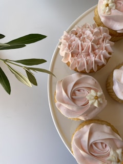 A few cupcakes with light pink frosting are arranged on a white plate. One cupcake is decorated with ruffled icing, while the others feature a swirl design and small white flower embellishments with yellow centers. A sprig of green leaves is visible beside the plate on a light background.