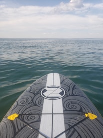 A paddleboard with a black and white swirling pattern on its deck is floating on calm, blue water. The horizon is visible in the distance under a partly cloudy sky.