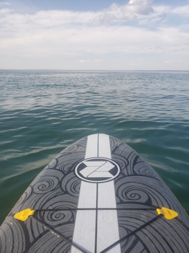 A paddleboard with a black and white swirling pattern on its deck is floating on calm, blue water. The horizon is visible in the distance under a partly cloudy sky.