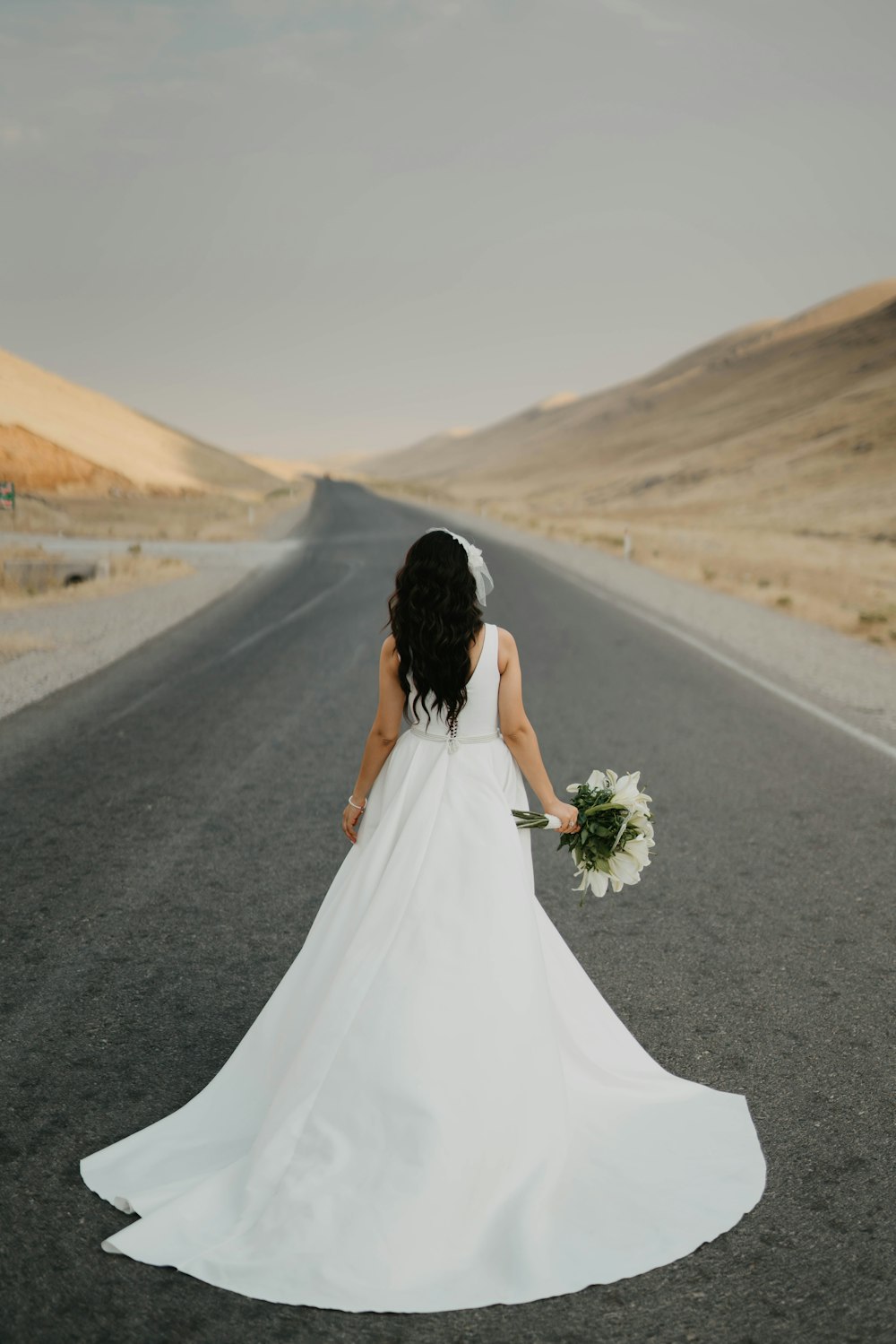 woman in white wedding dress holding bouquet of flowers walking on gray asphalt road during daytime