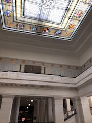 A grand interior space featuring a large stained glass ceiling with intricate designs, geometric patterns, and symbols. The walls are adorned with classical architectural elements, including ornate moldings and columns. The light is soft and diffused, creating a serene atmosphere.
