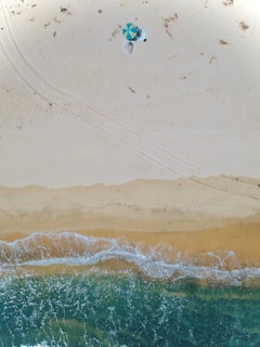 An aerial view of a sandy beach with clear turquoise water gently lapping at the shore. A small area of the beach has two people under a colorful umbrella with beach towels spread out next to them. The beach appears vast and mostly empty, with tracks and sparse vegetation visible on the sand.