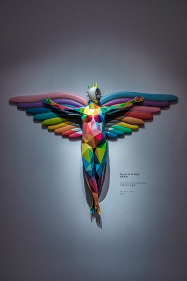 A vibrant, geometric sculpture with outstretched arms and colorful wings reminiscent of a bird or angel. The figure features a spiky crown on its head and a mask-like face, using a pattern of colorful polygons across its surface. The artwork is mounted on a plain wall with a spotlight casting a shadow and includes descriptive text alongside it.