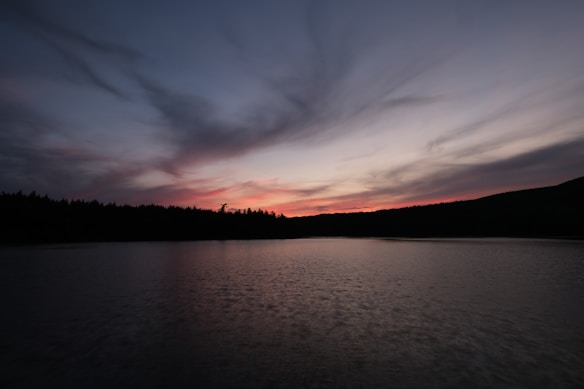 The image captures a serene lakeside view during twilight, with a silhouetted treeline along the horizon. The sky is painted in soft gradients of orange, pink, and purple with wispy clouds, reflecting beautifully on the water.
