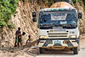 A truck labeled with Goldrock Construction is parked on a dirt road adjacent to a steep, vegetation-covered hill. Three workers are focused on constructing a wooden framework on the side of the road.