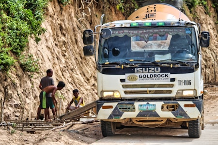 A truck labeled with Goldrock Construction is parked on a dirt road adjacent to a steep, vegetation-covered hill. Three workers are focused on constructing a wooden framework on the side of the road.