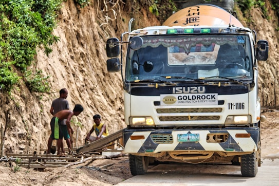 A truck labeled with Goldrock Construction is parked on a dirt road adjacent to a steep, vegetation-covered hill. Three workers are focused on constructing a wooden framework on the side of the road.