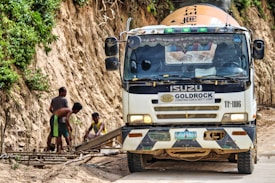 A truck labeled with Goldrock Construction is parked on a dirt road adjacent to a steep, vegetation-covered hill. Three workers are focused on constructing a wooden framework on the side of the road.