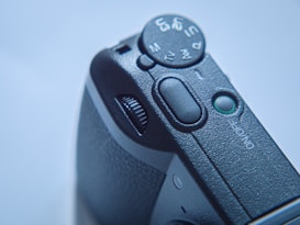A close-up view of a black camera dial displaying various settings, including TV, AV, and Manual. The textured surface of the camera body is visible, along with an on/off button and a small green light indicator.