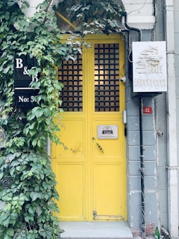 A vibrant yellow door with a grid pattern stands out against a muted facade. Lush green ivy partially covers the doorway, adding a natural element to the scene. A black sign with white lettering showing 'B & B No: 31' is mounted to the left. A certificate of excellence plaque and yellow tape with the word 'love' are fixed on the door. To the right, an electrical box with faded writing is visible. A small green bottle and some rust on the pipes add to the urban aesthetic.