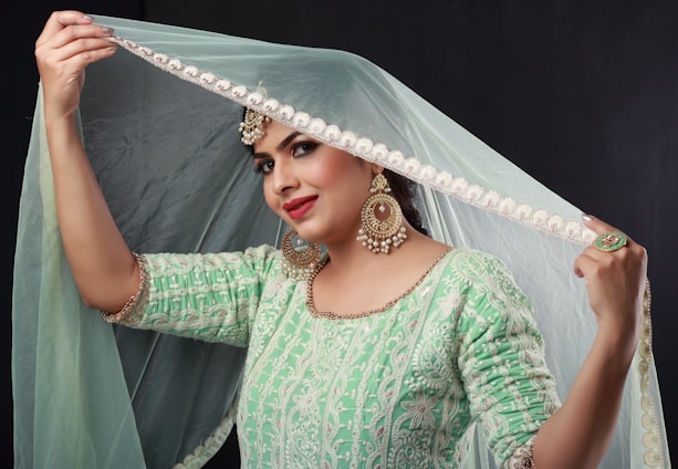 A woman wearing an ornate green dress looks to the side while holding up the edge of a delicate light blue dupatta adorned with embellishments. She is accessorized with large, decorative earrings, a ring, and a headpiece, and has a confident expression.