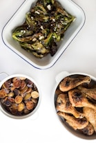 Three dishes are arranged on a white surface. One dish contains roasted Brussels sprouts sprinkled with cheese crumbles. Another bowl holds a mix of seasoned and roasted potato slices, garnished with small, purple flowers. A third dish features a stack of well-seasoned and roasted chicken drumsticks, showcasing golden brown skin.