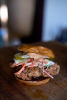 A close-up of a gourmet fried chicken sandwich featuring a piece of crispy fried chicken on a toasted bun. The sandwich is topped with slices of pickle, shredded carrots, coleslaw, and a drizzle of creamy sauce.