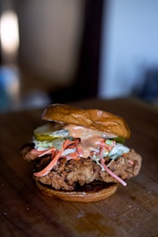 A close-up of a gourmet fried chicken sandwich featuring a piece of crispy fried chicken on a toasted bun. The sandwich is topped with slices of pickle, shredded carrots, coleslaw, and a drizzle of creamy sauce.