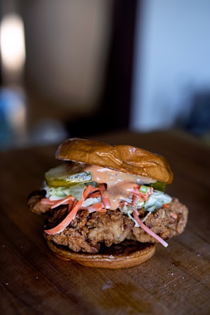 A close-up of a gourmet fried chicken sandwich featuring a piece of crispy fried chicken on a toasted bun. The sandwich is topped with slices of pickle, shredded carrots, coleslaw, and a drizzle of creamy sauce.