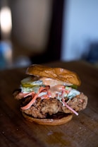 A close-up of a gourmet fried chicken sandwich featuring a piece of crispy fried chicken on a toasted bun. The sandwich is topped with slices of pickle, shredded carrots, coleslaw, and a drizzle of creamy sauce.