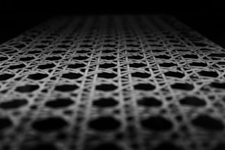 A close-up view of a woven pattern featuring interlacing lines forming hexagonal shapes. The lighting highlights the texture and depth of the weave against a dark background.