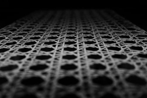 A close-up view of a woven pattern featuring interlacing lines forming hexagonal shapes. The lighting highlights the texture and depth of the weave against a dark background.