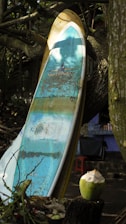 A weathered surfboard with blue and white colors is leaning against a tree. The surroundings include a partially opened coconut placed on a wooden surface and some foliage. A small, red stool and a building with a patterned cloth hanging can be seen in the background.