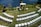 An outdoor wedding setup features neatly arranged white chairs facing a wooden arch adorned with white fabric and floral decorations. The setting overlooks a calm body of water, surrounded by lush greenery and a well-kept lawn.