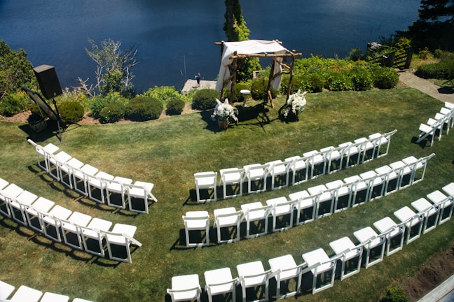 An outdoor wedding setup features neatly arranged white chairs facing a wooden arch adorned with white fabric and floral decorations. The setting overlooks a calm body of water, surrounded by lush greenery and a well-kept lawn.