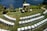 An outdoor wedding setup features neatly arranged white chairs facing a wooden arch adorned with white fabric and floral decorations. The setting overlooks a calm body of water, surrounded by lush greenery and a well-kept lawn.
