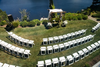 An outdoor wedding setup features neatly arranged white chairs facing a wooden arch adorned with white fabric and floral decorations. The setting overlooks a calm body of water, surrounded by lush greenery and a well-kept lawn.