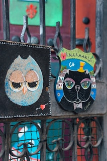 Two artistic signs are attached to an iron gate. The left sign depicts an owl with closed eyes, featuring a blue outer edge and a heart symbol at the bottom. The right sign showcases a black cat wearing a top hat and bow tie, set against a colorful village backdrop, with 'Marmaris' written at the top.