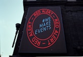 A large digital billboard on a brick building featuring a circular red border with the words 'RED ALERT' repeated twice. Inside the circle, the text '#WE MAKE EVENTS' is displayed in blue. The sign is illuminated against the backdrop of the building, conveying a clear message.