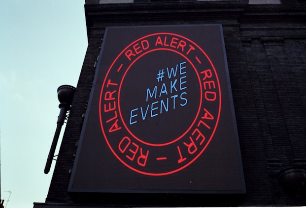 A large digital billboard on a brick building featuring a circular red border with the words 'RED ALERT' repeated twice. Inside the circle, the text '#WE MAKE EVENTS' is displayed in blue. The sign is illuminated against the backdrop of the building, conveying a clear message.