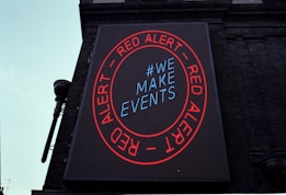 A large digital billboard on a brick building featuring a circular red border with the words 'RED ALERT' repeated twice. Inside the circle, the text '#WE MAKE EVENTS' is displayed in blue. The sign is illuminated against the backdrop of the building, conveying a clear message.