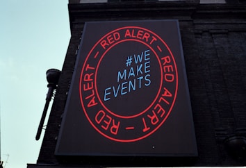 A large digital billboard on a brick building featuring a circular red border with the words 'RED ALERT' repeated twice. Inside the circle, the text '#WE MAKE EVENTS' is displayed in blue. The sign is illuminated against the backdrop of the building, conveying a clear message.