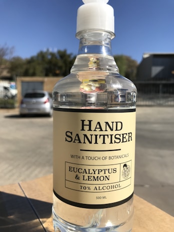 A clear plastic bottle of hand sanitizer with a white pump dispenser, labeled with 'Hand Sanitiser' and ingredients including eucalyptus and lemon. The bottle indicates it contains 70% alcohol and has a capacity of 500 ml. It is placed on an outdoor table with blurred background showing a parked car and trees under a clear blue sky.