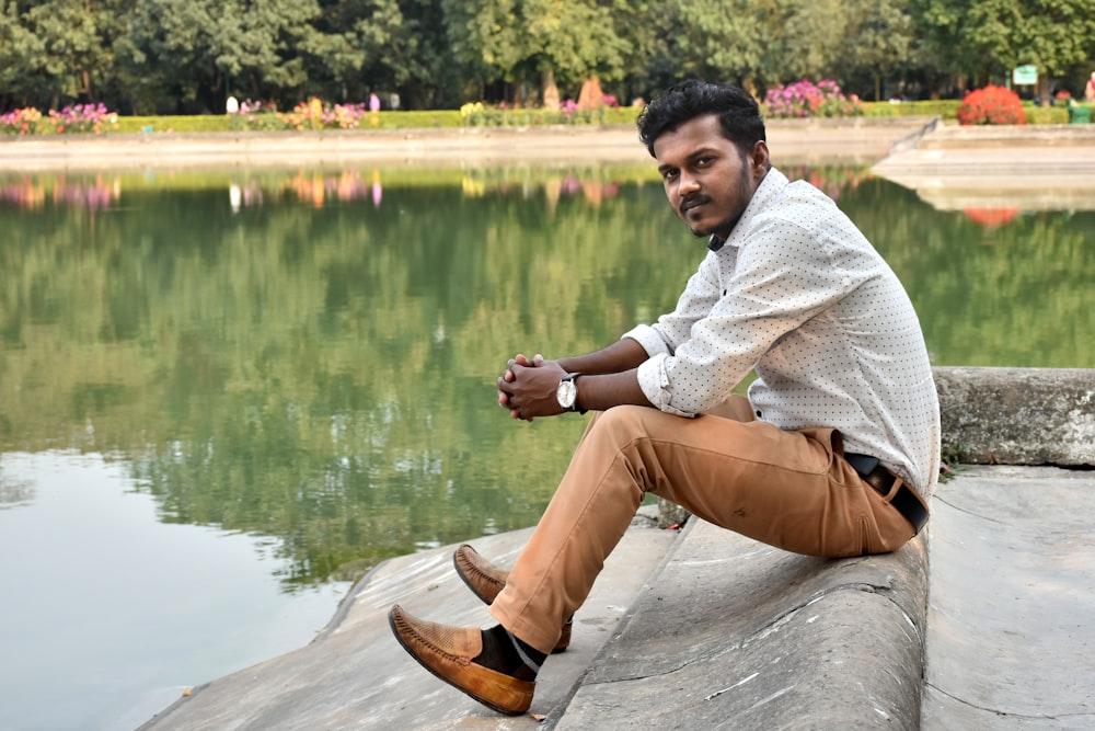 boy in white sweater and brown pants sitting on gray concrete surface near body of water