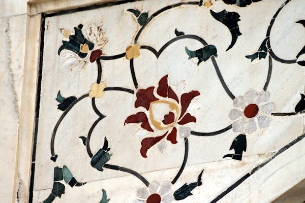 A detailed floral pattern inlaid into a marble surface, featuring red, white, and green leaf-like designs. The inlay uses intricately carved pieces to form a symmetrical arrangement around a central floral motif.