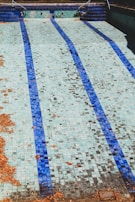 An empty swimming pool is depicted with blue and white tiled floor. There are scattered dry leaves across the pool, indicating it has not been in use for some time. The steps leading into the pool are visible, adding to the sense of abandonment.