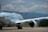 A large passenger airplane on a runway, featuring Air Canada livery. The aircraft is positioned with its rear and one wing visible, set against a backdrop of distant mountains and a cloudy sky. The tarmac is clear, and the environment suggests an airport or airfield with surrounding greenery.