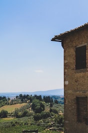 A rustic stone building is partially visible on the right side, overlooking a picturesque landscape of rolling hills and lush greenery. The sky is clear and blue, with cypress and other trees scattered across the countryside.