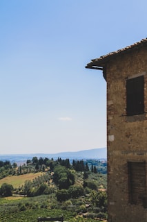 A rustic stone building is partially visible on the right side, overlooking a picturesque landscape of rolling hills and lush greenery. The sky is clear and blue, with cypress and other trees scattered across the countryside.