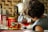A child with curly hair is pouring tea from a glass teapot into a transparent glass cup. On the counter, various tea boxes and a large red container are visible. The background features a tiled wall with a kettle nearby, suggesting a kitchen setting.