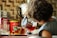 A child with curly hair is pouring tea from a glass teapot into a transparent glass cup. On the counter, various tea boxes and a large red container are visible. The background features a tiled wall with a kettle nearby, suggesting a kitchen setting.