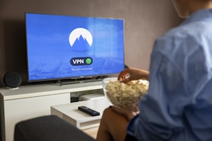 A person sits on a sofa facing a large flat-screen TV. The screen displays a VPN app interface indicating a secure connection. The person holds a bowl of popcorn and is dressed casually. A remote control lies on a white coffee table in front of them. The room is sparsely decorated with a sound speaker on the TV stand.