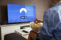A person sits on a sofa facing a large flat-screen TV. The screen displays a VPN app interface indicating a secure connection. The person holds a bowl of popcorn and is dressed casually. A remote control lies on a white coffee table in front of them. The room is sparsely decorated with a sound speaker on the TV stand.