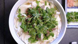 A bowl of noodle soup topped with fresh herbs like cilantro or basil sits on a table. Large pieces of white onion and slices of beef are visible beneath the herbs in the broth. In the background, there are small side dishes containing pickled ginger and additional herbs.