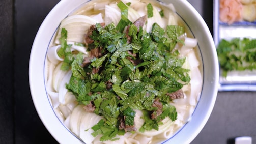 A bowl of noodle soup topped with fresh herbs like cilantro or basil sits on a table. Large pieces of white onion and slices of beef are visible beneath the herbs in the broth. In the background, there are small side dishes containing pickled ginger and additional herbs.