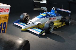 A vintage Formula One racing car is displayed indoors. The car features a sleek aerodynamic design with prominent sponsor logos, including Toshiba and Castrol, on its blue, white, and yellow painted body. The tires are robust, emphasizing the vehicle’s readiness for high-speed racing.