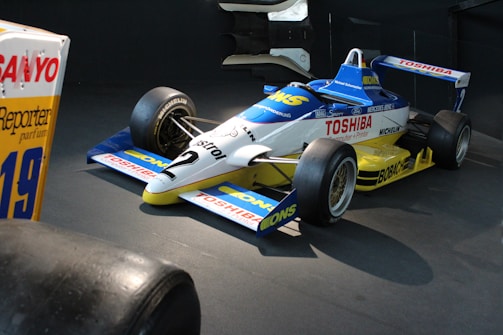 A vintage Formula One racing car is displayed indoors. The car features a sleek aerodynamic design with prominent sponsor logos, including Toshiba and Castrol, on its blue, white, and yellow painted body. The tires are robust, emphasizing the vehicle’s readiness for high-speed racing.