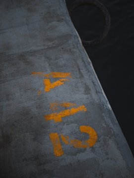 Concrete surface with weathered yellow numbers painted on it, shown in an industrial environment with a dark body of water to the side.